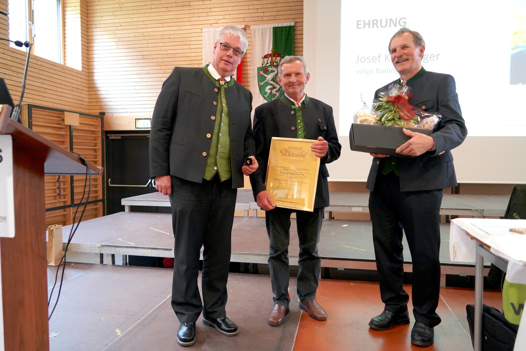 Drei Männer bei einer Ehrung mit Urkunde und Geschenk auf einer Bühne