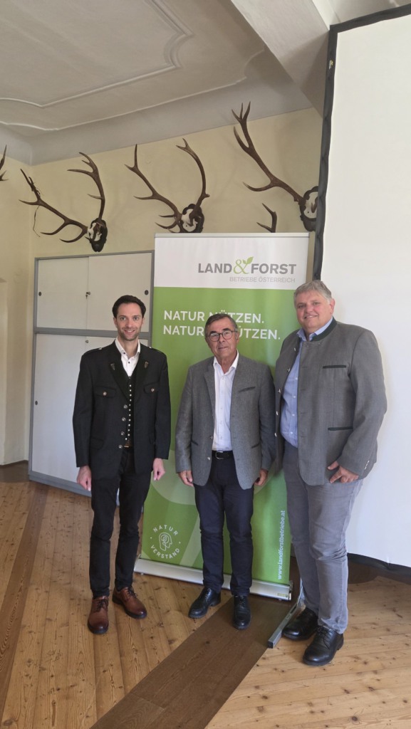 Drei Männer vor Banner von Land & Forst Betriebe Österreich, darüber Geweihe an der Wand